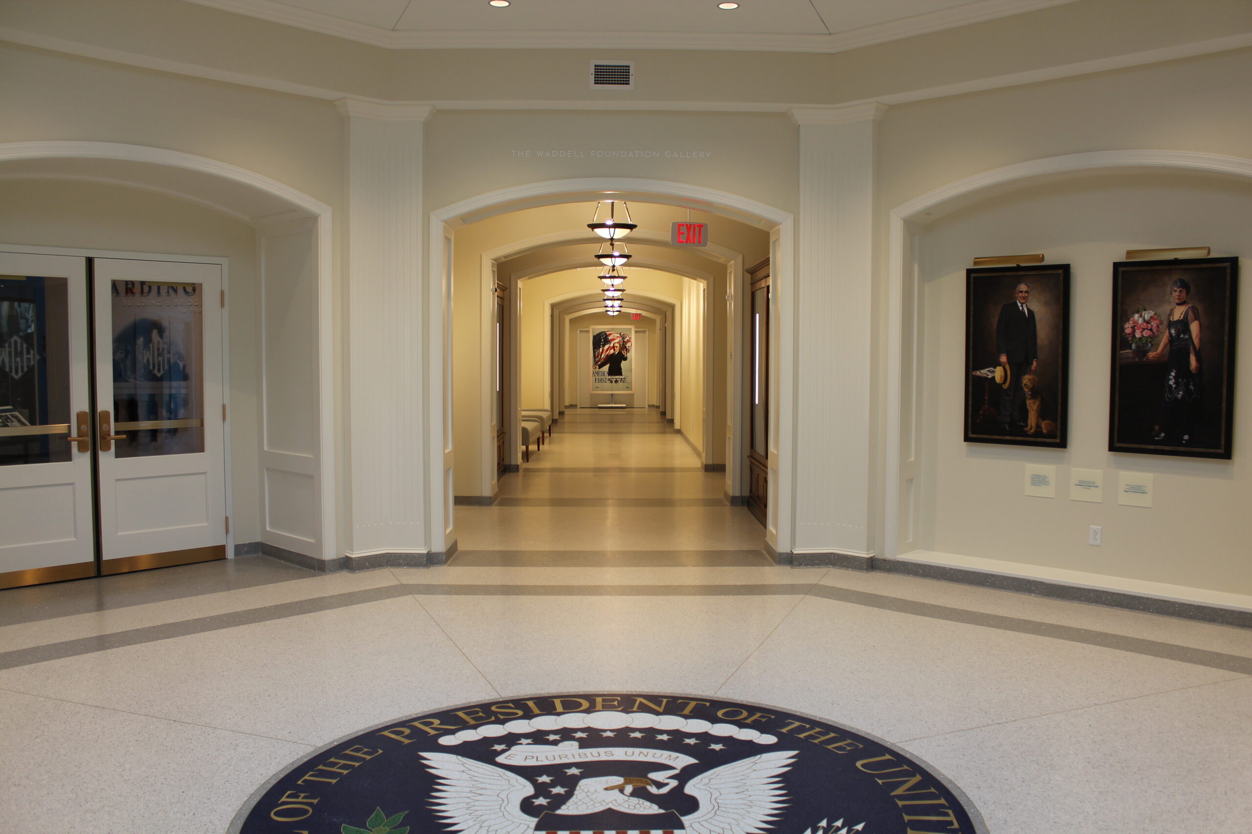 Corridor within the Harding Presidential Library & Museum with doors to the left leading to exhibit space and portraits of Florence and Warren Harding on the right. A Presidential seal sits in the middle of the room.