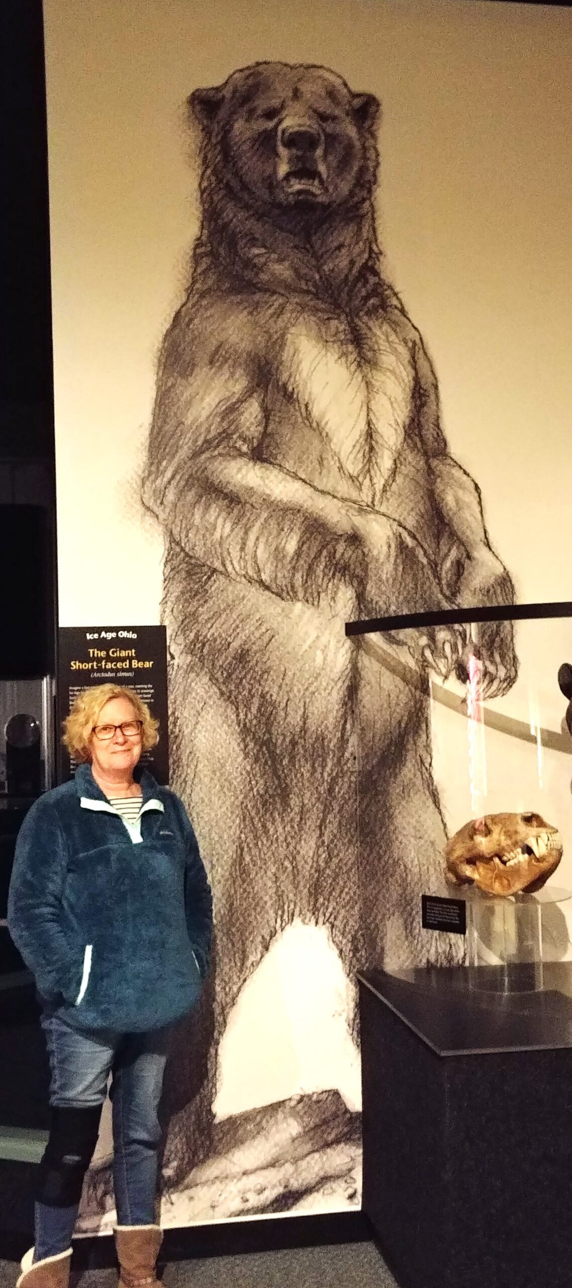 A woman standing next to a life-size illustration of a Giant Short-faced Bear standing upright on its hind legs.
